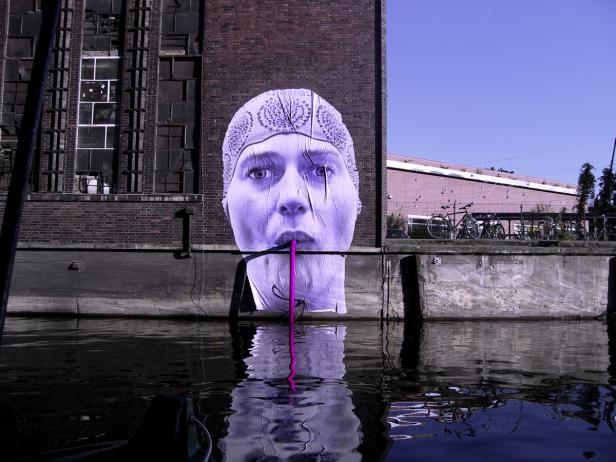 Ein großes Porträt einer Frau mit Kopftuch an einer Backsteinmauer am Wasser.