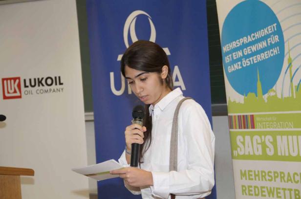 Eine junge Frau hält ein Mikrofon und liest von einem Blatt Papier vor Bannern von Lukoil und „Sag's Multi“.