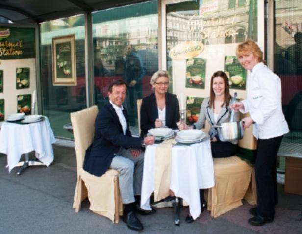 Knorr - Out-of-Home-Kampagne - Knorr Gourmet Suppen - Mindshare Austria - Gewista Drei Personen sitzen an einem gedeckten Tisch, während eine Köchin Suppe serviert.