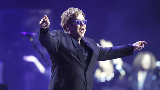 Elton John tritt in einem schwarzen Anzug und blauen Gläsern auf einer Bühne auf.