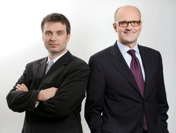 Zwei Männer in Anzügen stehen nebeneinander vor einem hellen Hintergrund.