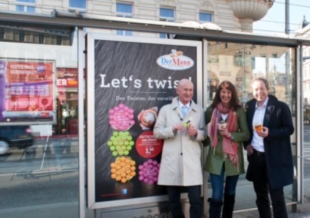 Drei Personen stehen vor einer Bushaltestelle mit Werbung für „Der Mann“-Süßwaren.