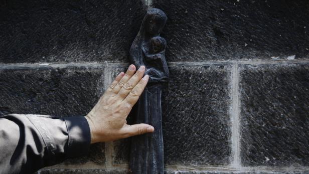 Eine Hand berührt eine Skulptur der Jungfrau Maria mit Kind an einer dunklen Steinmauer.