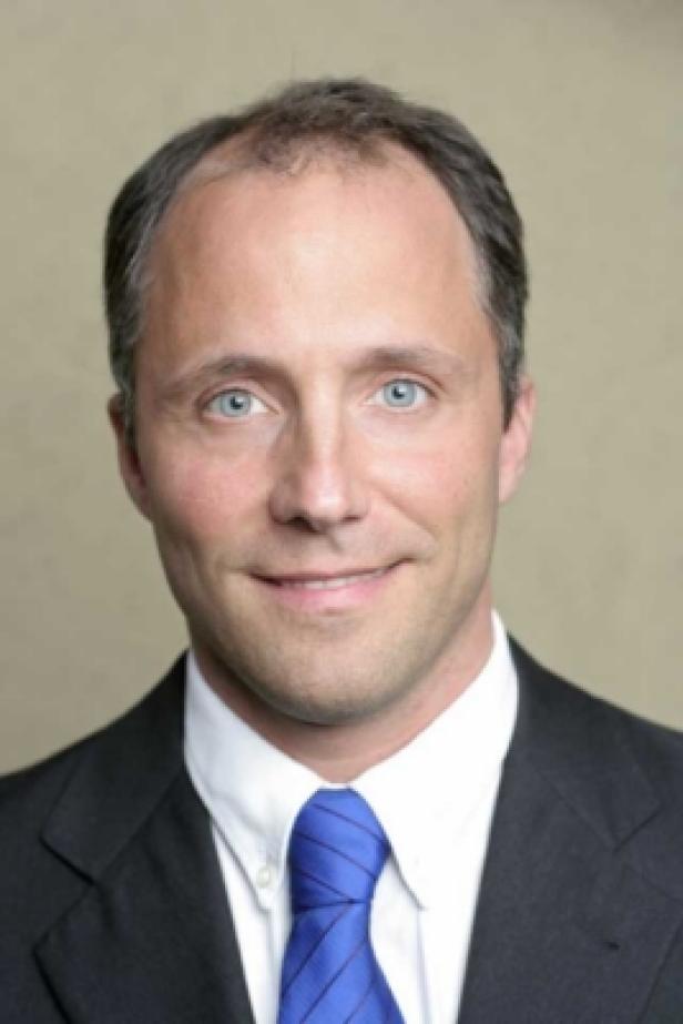 Ein Mann mit blauen Augen trägt einen dunklen Anzug und eine blaue Krawatte.