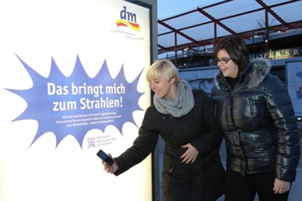 dm - Außenwerbe-Kampagne - Das bringt mich zum Strahlen - Epamedia - City Light (c: dm) Zwei Frauen betrachten eine DM-Werbung mit dem Slogan „Das bringt mich zum Strahlen!".