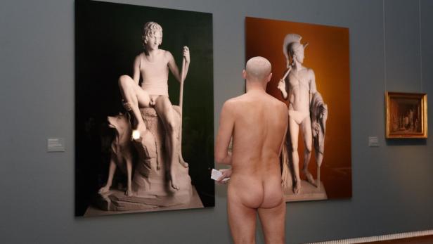 Ein nackter Mann betrachtet in einer Galerie zwei großformatige Bilder von Statuen.