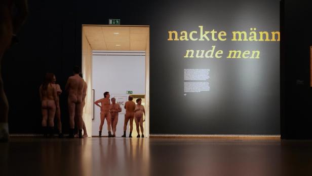 Eine Gruppe nackter Menschen steht in einem Museum vor der Aufschrift „nackte männer nude men“.
