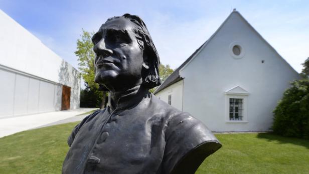 Eine Bronzebüste vor einem weißen Gebäude unter blauem Himmel.