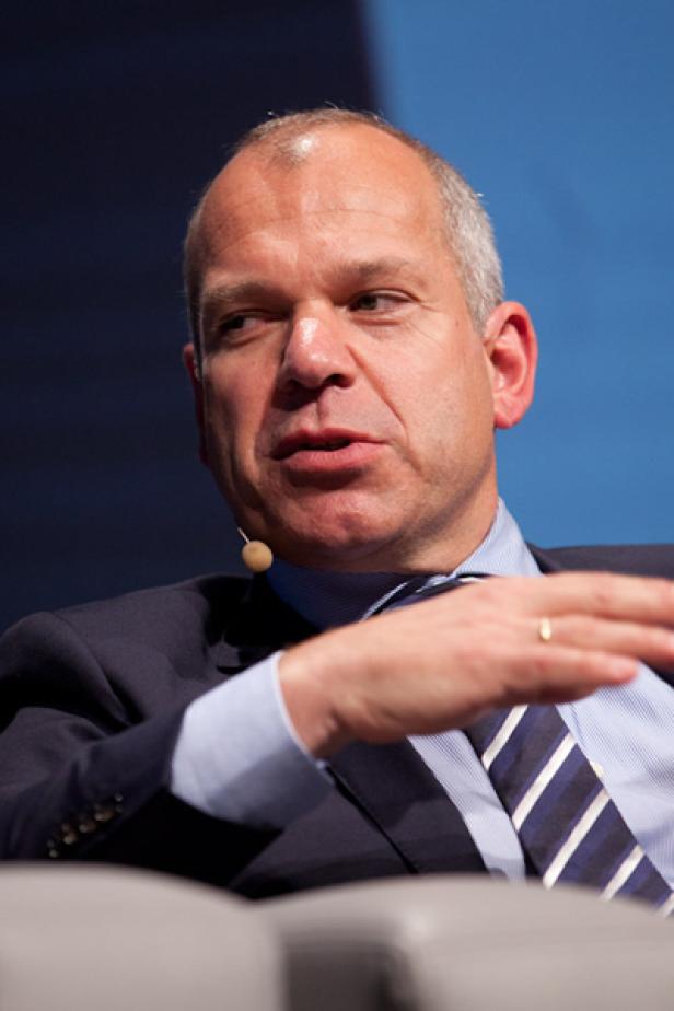 Ein Mann in Anzug und Krawatte spricht vor einem blauen Hintergrund.