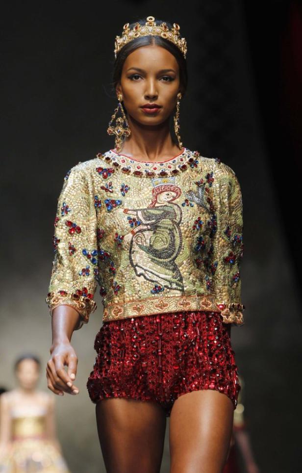 Ein Model mit Krone und Ohrringen präsentiert ein goldenes Oberteil und rote Shorts auf dem Laufsteg.