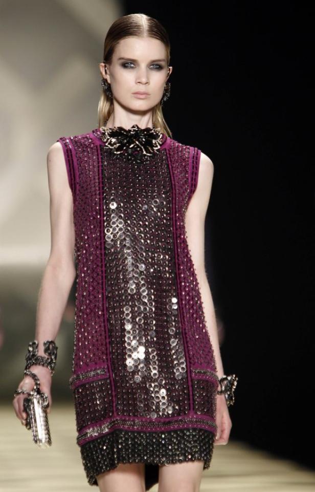 Ein Model präsentiert ein ärmelloses, lila Kleid mit silbernen Pailletten und dunklem Blumenschmuck am Hals.