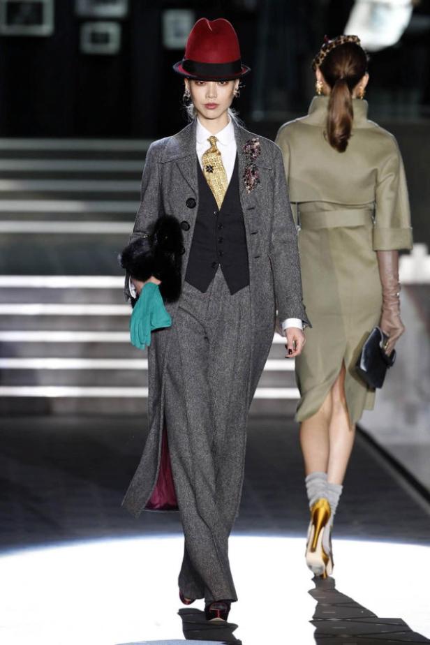Eine Frau in einem grauen Hosenanzug und einem roten Hut auf einem Laufsteg.