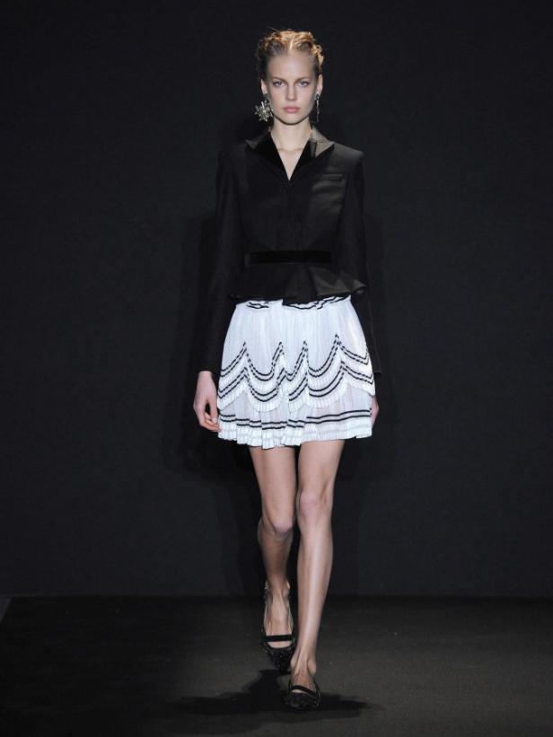 Ein Model präsentiert ein Outfit mit schwarzem Blazer und weißem, kurzem Rock auf dem Laufsteg.