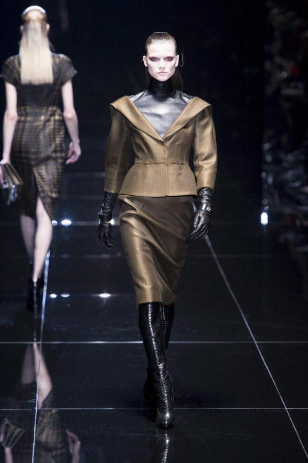 Eine Frau in einem goldenen Kostüm mit Blazer, Rock, schwarzen Handschuhen und Schlangenlederstiefeln auf dem Laufsteg.