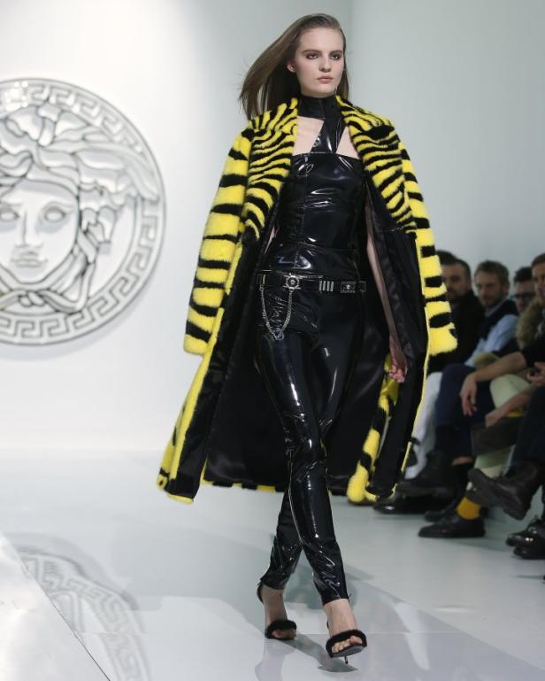 Ein Model in einem gelb-schwarz gestreiften Mantel und glänzender schwarzer Kleidung auf dem Laufsteg.