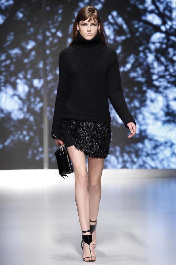 Eine Frau in einem schwarzen Rollkragenpullover und einem Fransenrock auf einem Laufsteg.