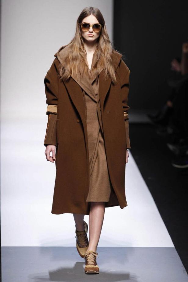 Eine Frau mit Sonnenbrille trägt einen braunen Mantel und ein Kleid auf dem Laufsteg.
