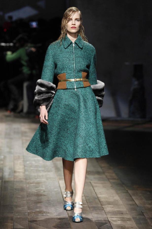 Ein Model präsentiert ein grünes Tweed-Kleid mit Pelzärmeln und goldenem Gürtel auf dem Laufsteg.