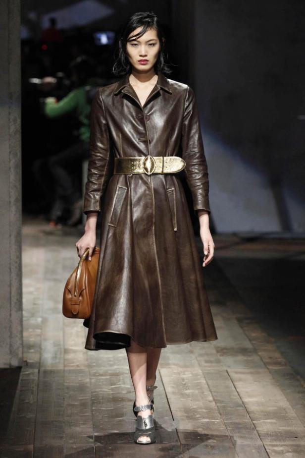 Eine Frau in einem braunen Ledermantel mit goldenem Gürtel und einer Handtasche auf einem Laufsteg.