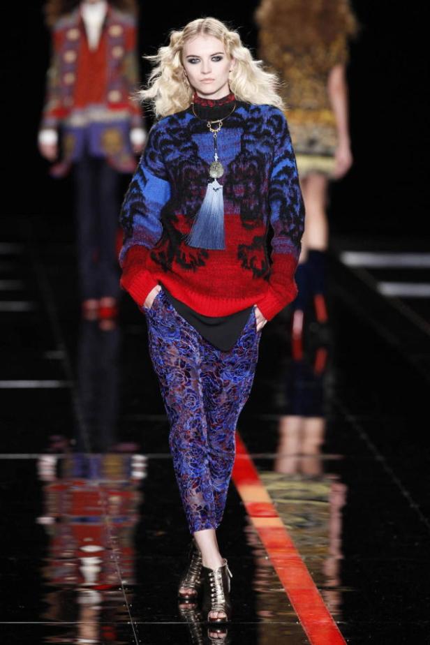 Ein blondes Model präsentiert einen roten Pullover mit blau-schwarzem Muster und blaue Hosen auf dem Laufsteg.