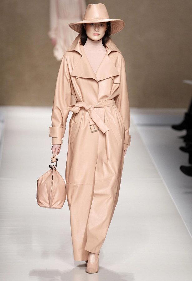 Eine Frau in einem hellrosa Trenchcoat und Hut auf einem Laufsteg.