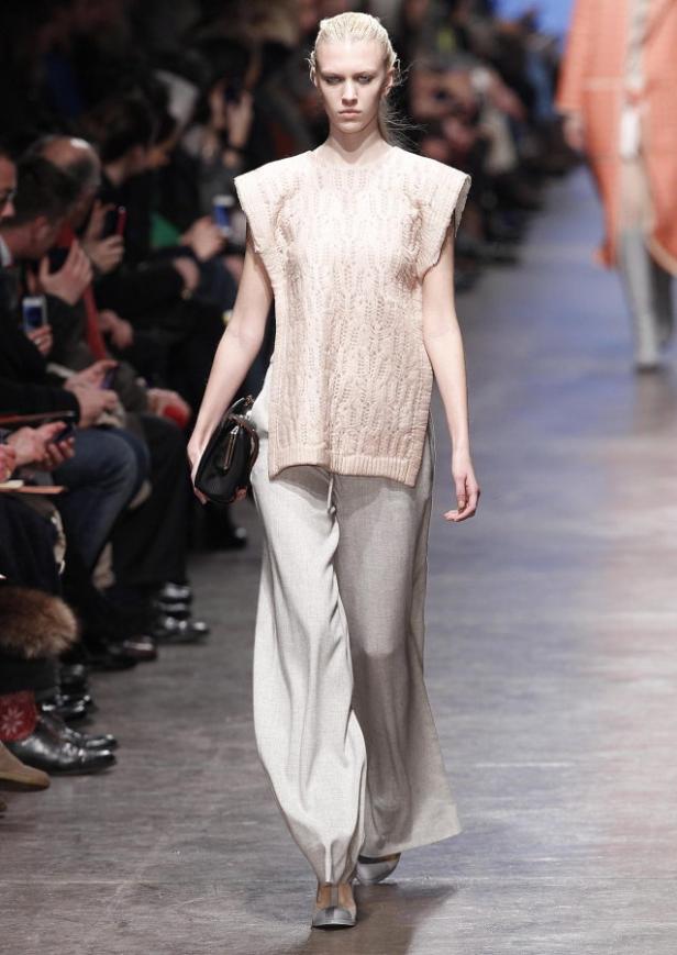 Eine Frau in einem hellrosa Strickoberteil und einer weiten, grauen Hose auf einem Laufsteg.