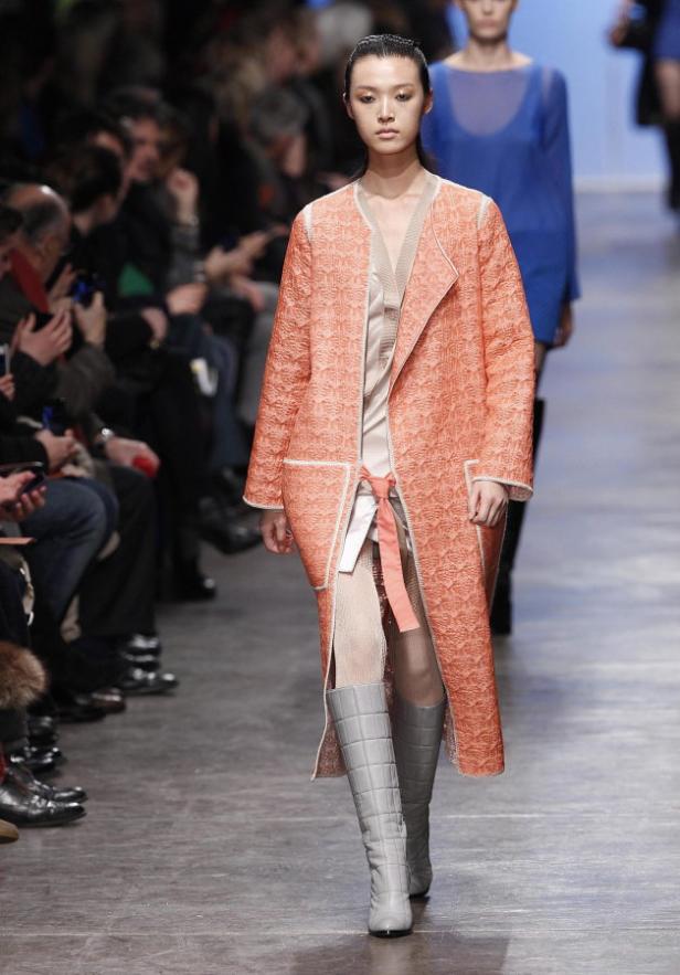Eine Frau auf dem Laufsteg trägt einen langen, gemusterten, lachsfarbenen Mantel und graue Stiefel.