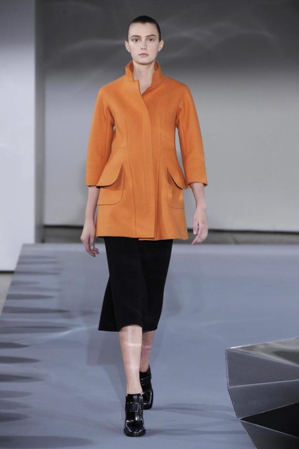 Eine Frau präsentiert einen orangefarbenen Mantel und einen schwarzen Rock auf einem Laufsteg.