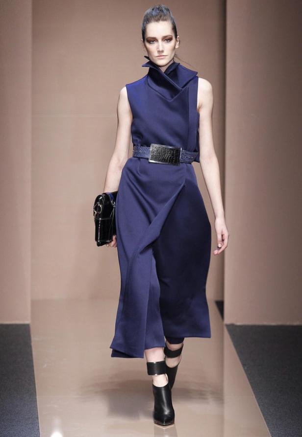 Eine Frau auf dem Laufsteg präsentiert ein blaues, ärmelloses Kleid mit breitem Gürtel und schwarzen Stiefeletten.