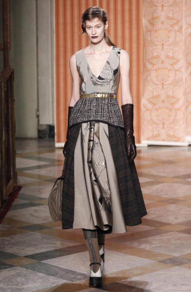 Eine Frau präsentiert ein ärmelloses Kleid mit goldenem Gürtel und braunen Handschuhen auf einem Laufsteg.