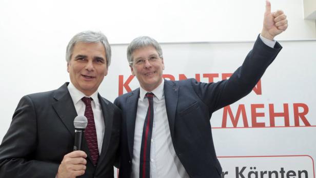 Zwei Männer in Anzügen posieren vor einem Banner mit der Aufschrift „Kärnten MEHR“.