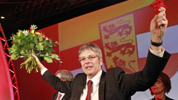 Ein Mann im Anzug hält einen Blumenstrauß und einen roten Gegenstand in die Höhe.