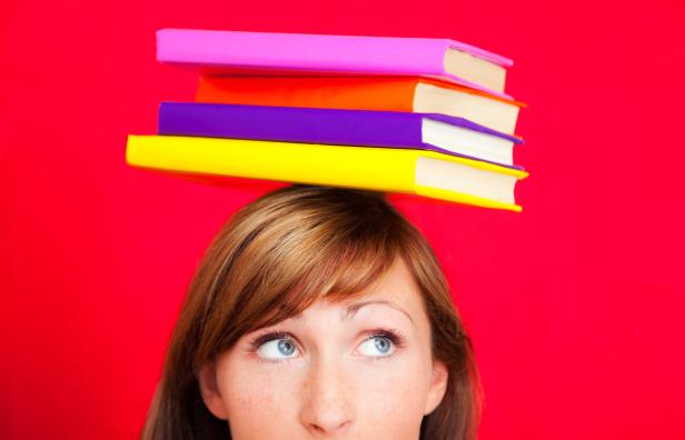 Eine Frau balanciert einen Stapel bunter Bücher auf ihrem Kopf vor einem roten Hintergrund.