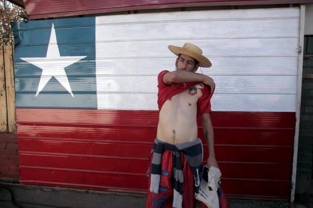 Ein Mann mit Hut und rotem T-Shirt steht vor einer Wand, die wie die chilenische Flagge bemalt ist.