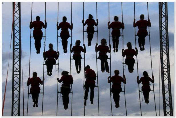 Eine Gruppe von Personen in roter Kleidung sitzt auf Hängeleitern vor einem bewölkten Himmel.