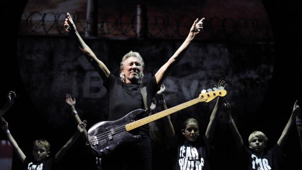 Roger Waters mit erhobenen Armen und Bassgitarre auf der Bühne, umgeben von Kindern.