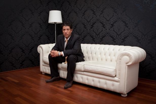 Paul Klitsch, Senior Consultant Home Gruppe (c: home gruppe) Ein Mann im Anzug sitzt auf einem weißen Sofa vor einer dunklen Wand.