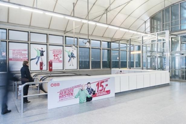 Eine Frau wartet an einer Rolltreppe in einer ÖBB-Station mit Werbung für Tickets „Ab in den Westen“.