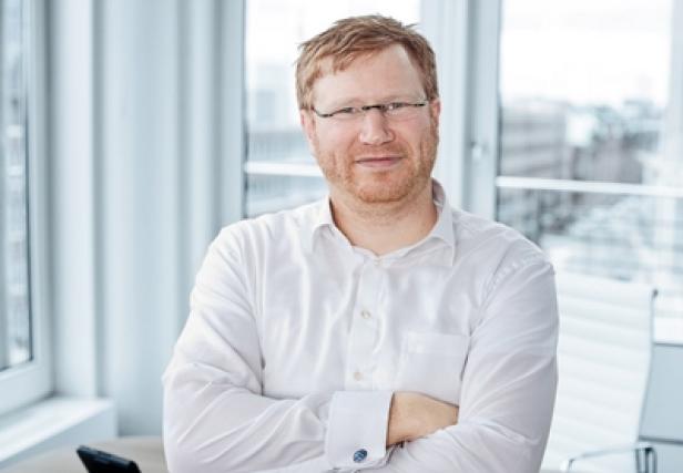 Ein Mann mit Brille und Hemd steht mit verschränkten Armen vor einem Fenster.