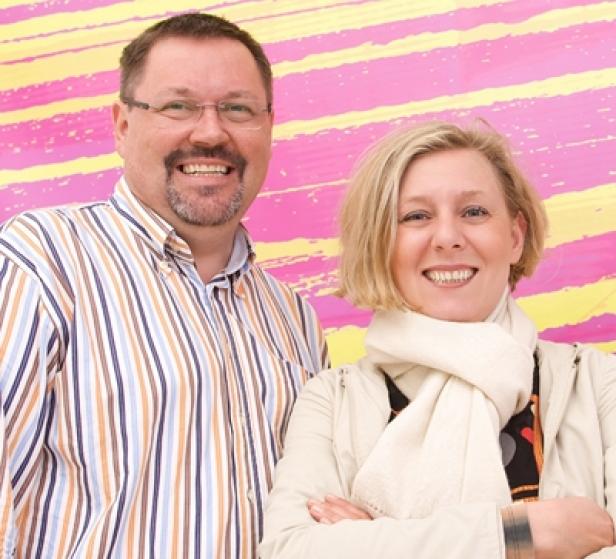 Ein Mann mit Brille und eine Frau mit Schal lächeln vor einem farbenfrohen Hintergrund.