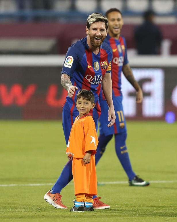 Lionel Messi mit einem Kind auf dem Fußballfeld, Neymar im Hintergrund.