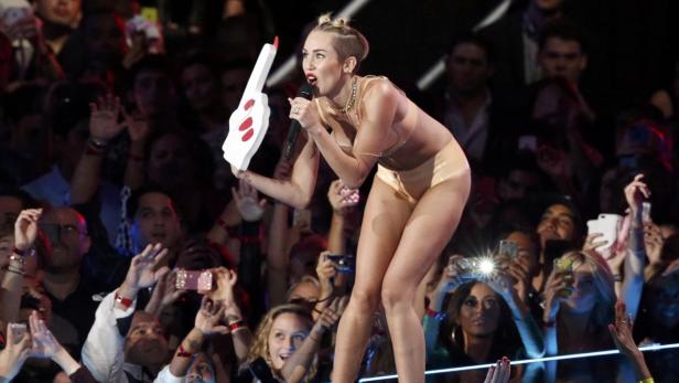 Miley Cyrus tritt mit einem Mikrofon und einem übergroßen Schaumstoffhandschuh auf einer Bühne auf.