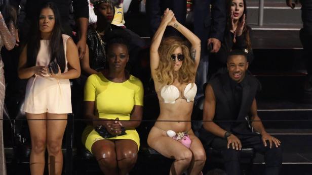 Lady Gaga in einem Muschel-BH bei einer Veranstaltung, umgeben von anderen Zuschauern.