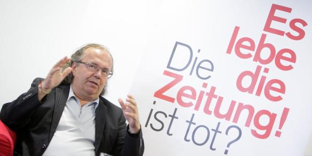 Ein Mann gestikuliert vor einem Plakat mit der Aufschrift „Die Zeitung ist tot? Es lebe die Zeitung!“.