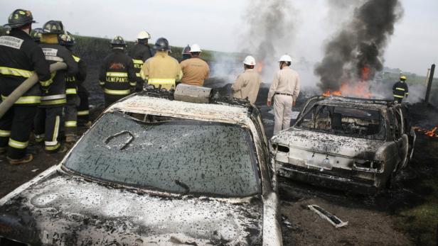 Feuerwehrleute stehen neben zwei ausgebrannten Autos, während im Hintergrund Rauch aufsteigt.