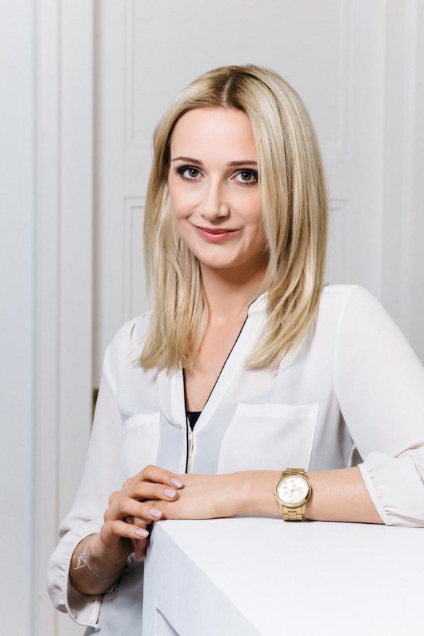 Eine blonde Frau mit einem Lächeln und einer goldenen Uhr posiert vor einer weißen Tür.