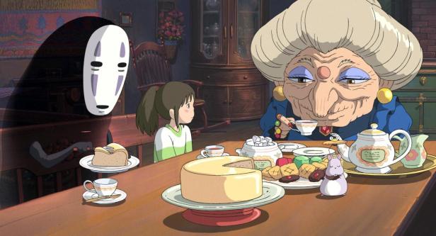Chihiro sitzt mit No-Face und Yubaba an einem Tisch voller Speisen in „Chihiros Reise ins Zauberland“.