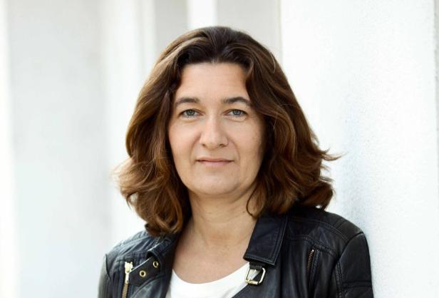Angelika Kramer Eine Frau mit braunen Haaren und einer schwarzen Lederjacke lehnt an einer weißen Wand.