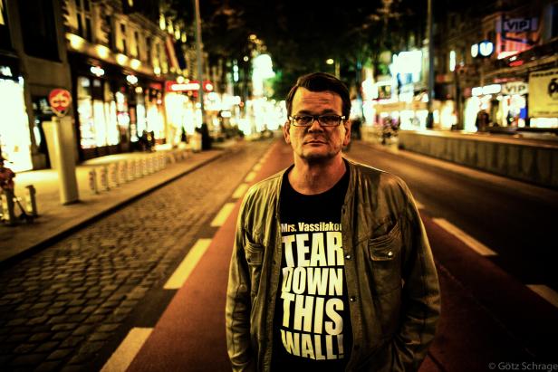 Ein Mann mit Brille und T-Shirt mit der Aufschrift „Tear Down This Wall“ steht nachts auf einer belebten Straße.