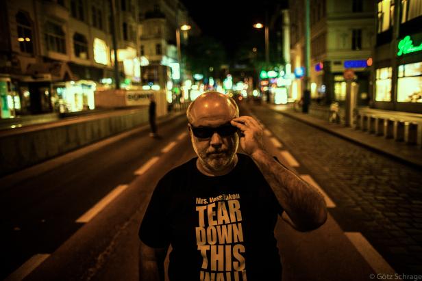 Ein Mann mit Sonnenbrille und einem T-Shirt mit der Aufschrift „Tear Down This Wall“ steht nachts auf einer Straße.